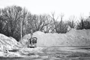 A black and white photograph of an excavator clearing snow from a parking lot--not quite the same as winter sweeping, but a nice image.